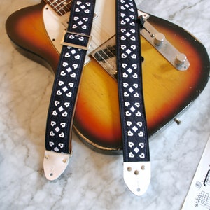 May include: Two black guitar straps with white heart-shaped patterns. The straps are lying on top of a sunburst electric guitar.