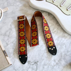 Könnte beinhalten: Ein roter, gelber und schwarzer Gitarrengurt mit einem Blumenmuster. Der Gurt hat eine braune Lederunterseite und schwarze Lederenden mit Metallnieten.