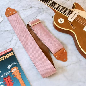 Op de afbeelding: Een roze corduroy gitaarband met bruine leren uiteinden en een zilveren gesp ligt naast een bruine elektrische gitaar. Een boek getiteld "Guitar Identification" en enkele plectrums zijn ook zichtbaar.