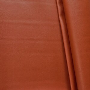 Könnte beinhalten: Nahaufnahme eines Stücks braunen Kunstleders. Der Stoff hat eine strukturierte Oberfläche und ist in der Mitte gefaltet.