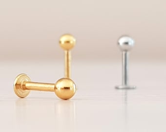 Puede incluir: Dos pendientes de barra de oro y uno de plata con bolas redondas en cada extremo. Los pendientes están sobre una superficie blanca.