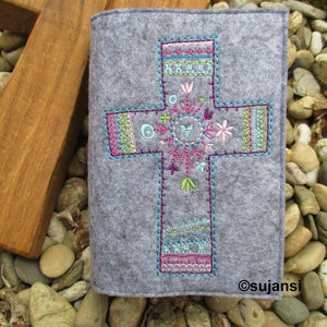 Könnte beinhalten: Ein grauer Filz-Buchbezug in Kreuzform mit bunter Stickerei. Die Stickerei zeigt ein Kreuz mit Blumen, Blättern und geometrischen Mustern in Rosa, Blau, Grün und Weiß.