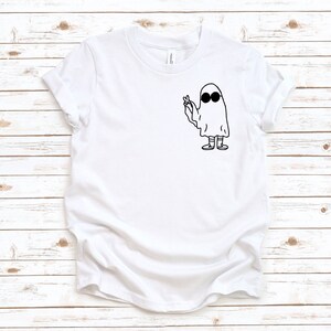 May include: White t-shirt featuring a black and white graphic of a ghost wearing sunglasses and giving a peace sign. The ghost is drawn in a simple, cartoon style. The shirt has short sleeves and a crew neck.