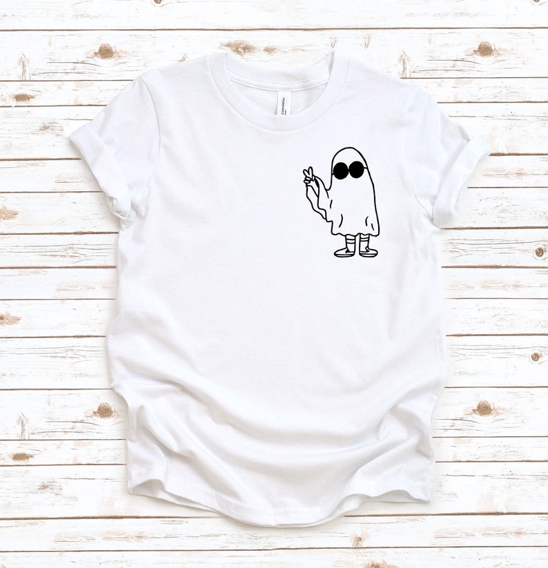 May include: White t-shirt featuring a black and white graphic of a ghost wearing sunglasses and giving a peace sign. The ghost is drawn in a simple, cartoon style. The shirt has short sleeves and a crew neck.