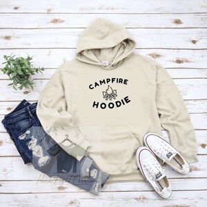 May include: A beige hoodie with the text "CAMPFIRE HOODIE" in black, along with a graphic of a campfire. The hoodie is paired with blue jeans and white sneakers. A small green plant is also in the image.
