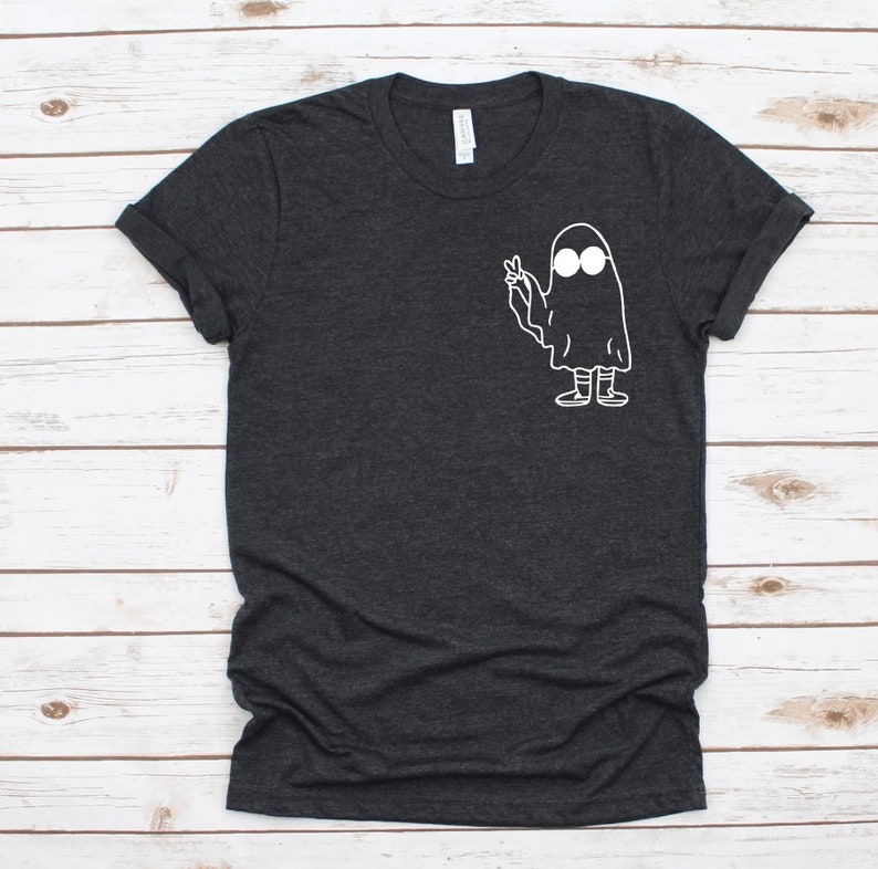 May include: Dark gray t-shirt featuring a white cartoon ghost with sunglasses making a peace sign. The ghost has a simple, minimalist design. The shirt has a crew neck and short sleeves.