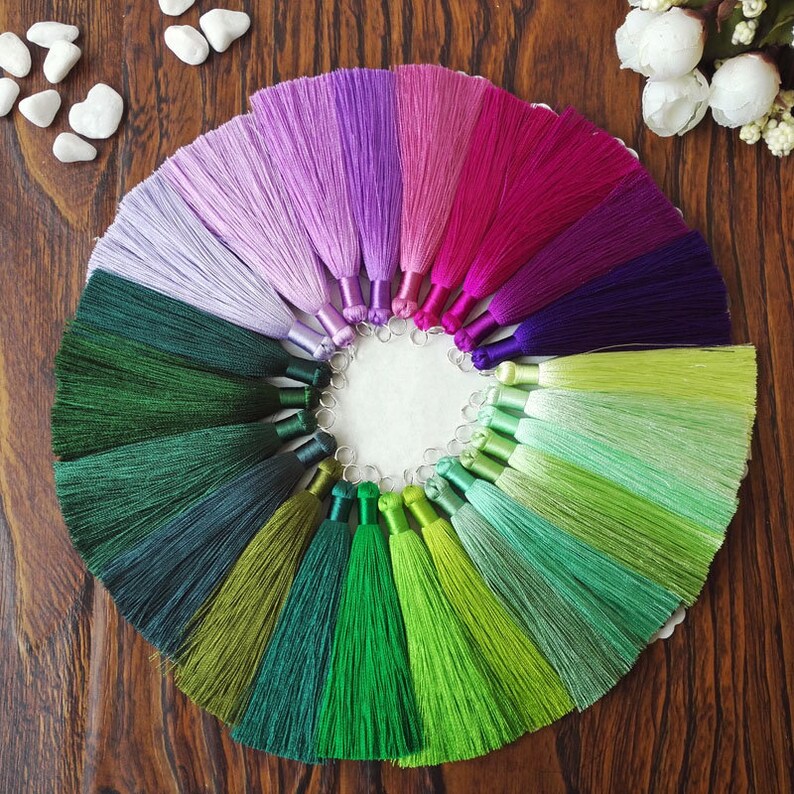 May include: A collection of 20 colorful silk tassels arranged in a circle on a wooden surface. The tassels are in shades of purple, pink, green, and blue.