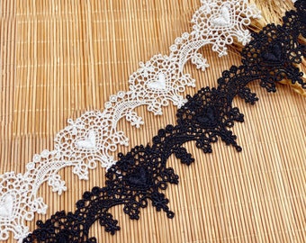 May include: Two lace trims, one black and one white, with a scalloped edge and a heart design. The trims are laid out on a woven bamboo surface.