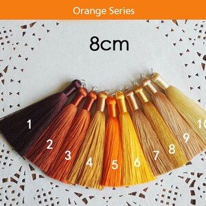 May include: Ten 8cm long tassels in shades of orange, yellow, and brown. The tassels are arranged in a semi-circle on a white lace doily. Each tassel is numbered 1 through 10.