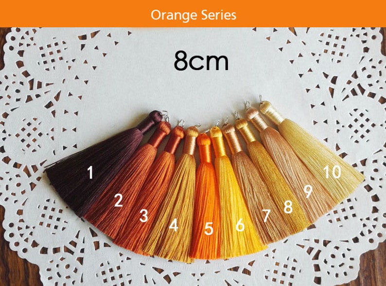 May include: Ten 8cm long tassels in shades of orange, yellow, and brown. The tassels are arranged in a semi-circle on a white lace doily. Each tassel is numbered 1 through 10.
