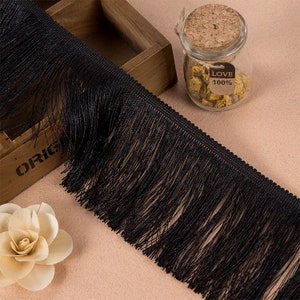 May include: A long strip of black fringe trim featuring a woven header and thin, dangling threads. The trim is displayed alongside a wooden box, a small glass jar with a cork stopper containing dried flowers and a label reading LOVE 100%.