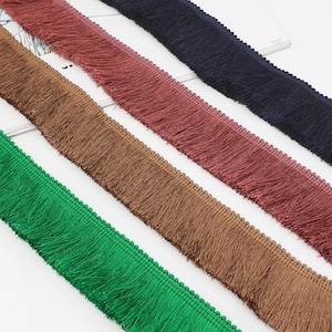 May include: Four strips of fringe trim arranged diagonally, featuring distinct colours including vibrant green, medium brown, deep maroon, and dark navy blue. Each strip has a woven header with a small beaded edge and long, straight tassels.