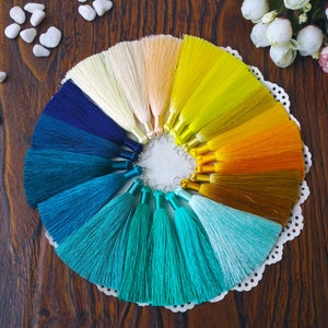 May include: A collection of 15 colorful silk tassels arranged in a circle on a white doily. The tassels range in color from white to yellow, orange, and blue.