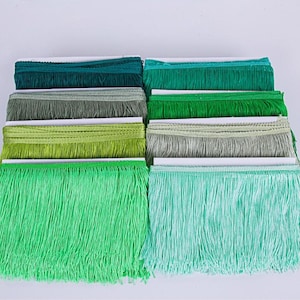 Puede incluir: Una variedad de adornos de flecos verdes en varios tonos, desde verde oscuro hasta menta claro. Los flecos están cuidadosamente apilados, mostrando las diferentes texturas y tonalidades del material decorativo.