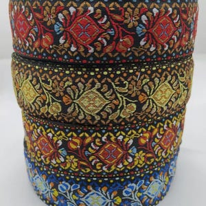 Op de afbeelding: Een stapel van vier rollen zwarte stoffen linten met complexe geweven patronen. Elke rol toont een eigen ontwerp met kleurrijk borduurwerk in tinten rood, goud, geel en blauw, met herhalende florale en geometrische motieven.