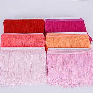 May include: A selection of colourful fringe trim in shades of red, pink, and orange. The fringe is neatly arranged in stacks, showcasing the variety of colours and textures. Ideal for craft projects, the trim adds a decorative element to clothing, accessories, and home decor.
