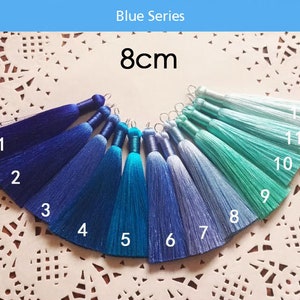 May include: A set of 12 blue and turquoise silk tassels, each measuring 8 cm in length. The tassels are arranged in a semi-circle on a white lace doily. The tassels are numbered 1 through 12.