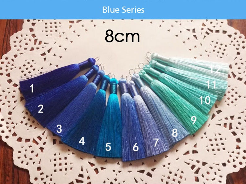 May include: A set of 12 blue and turquoise silk tassels, each measuring 8 cm in length. The tassels are arranged in a semi-circle on a white lace doily. The tassels are numbered 1 through 12.