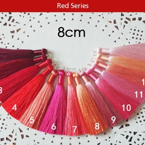 May include: A set of 14 tassel trims in various shades of red, pink, and orange. The tassels are 8 cm long and are arranged in a semi-circle on a white background.