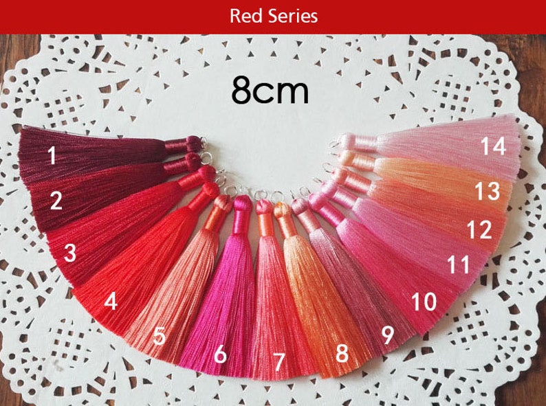 May include: A set of 14 tassel trims in various shades of red, pink, and orange. The tassels are 8 cm long and are arranged in a semi-circle on a white background.
