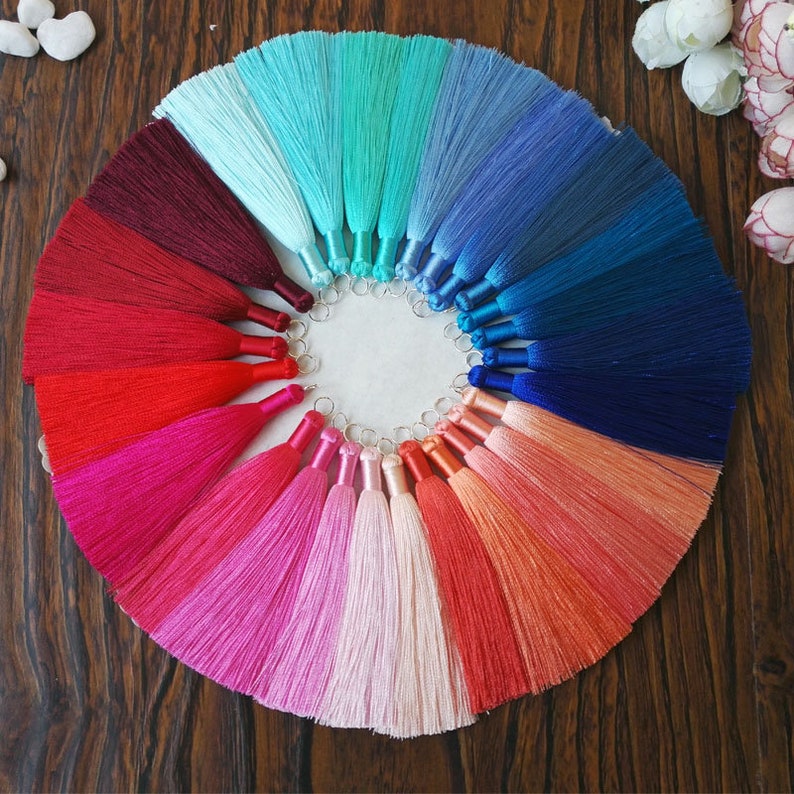 May include: A collection of 18 colorful silk tassels arranged in a circle on a wooden surface. The tassels are in shades of blue, green, red, pink, and orange.