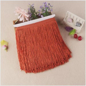 May include: A long, rust-orange fringe trim featuring a woven header band. The dense, straight strands hang vertically from the top edge, creating a uniform texture. The trim is displayed with a light pink flower and purple floral sprigs.