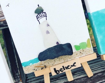 May include: A small white canvas painting of a lighthouse made from sea glass on a miniature easel. The easel has a sign that reads "believe".