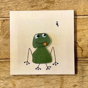 Peut inclure: Une grenouille verte avec des yeux globuleux et la langue tirée, faite de pierres peintes, sur un fond blanc.
