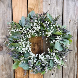 May include: A green and white wreath made of eucalyptus and baby's breath. The wreath is hanging on a wooden wall.