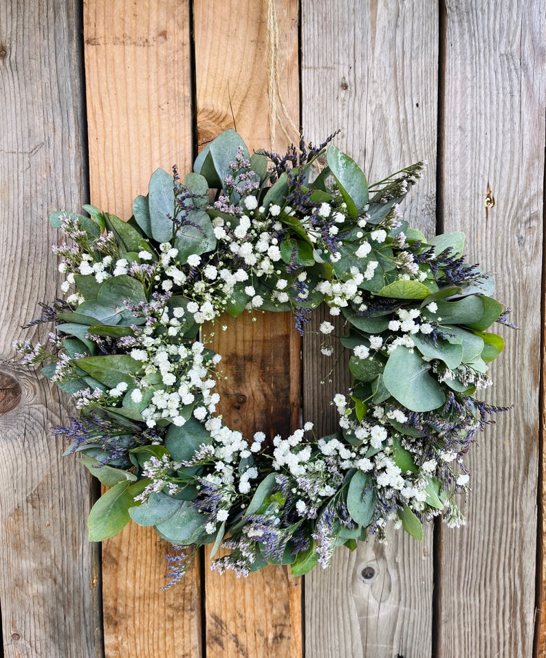 May include: A green and white wreath made of eucalyptus and baby's breath. The wreath is hanging on a wooden wall.