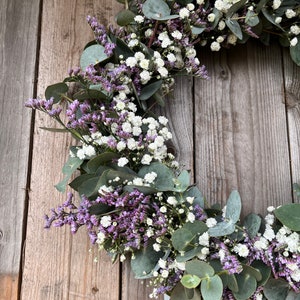 May include: A wreath made of eucalyptus leaves and small white and purple flowers. The wreath is hanging on a wooden background.