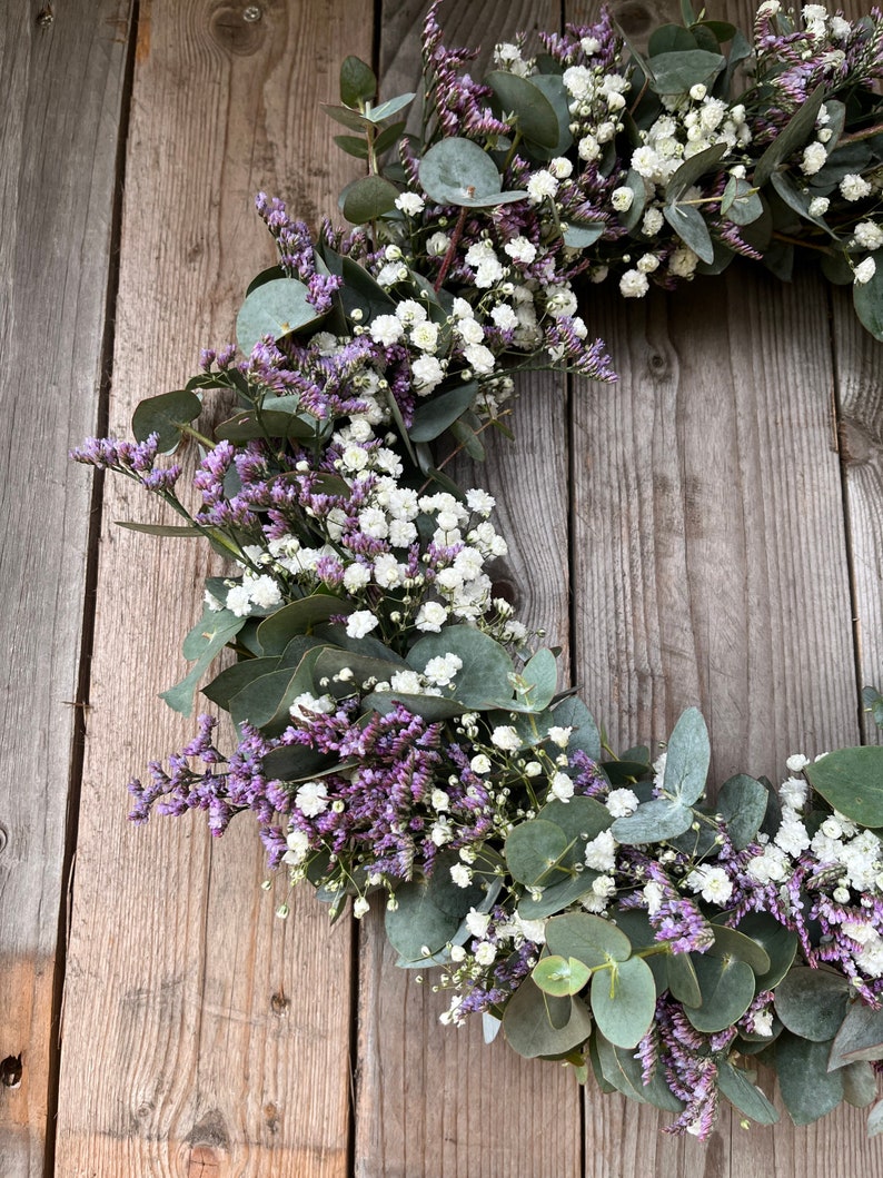 May include: A wreath made of eucalyptus leaves and small white and purple flowers. The wreath is hanging on a wooden background.