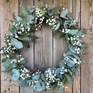 May include: A green eucalyptus and baby's breath wreath hanging on a wooden fence.
