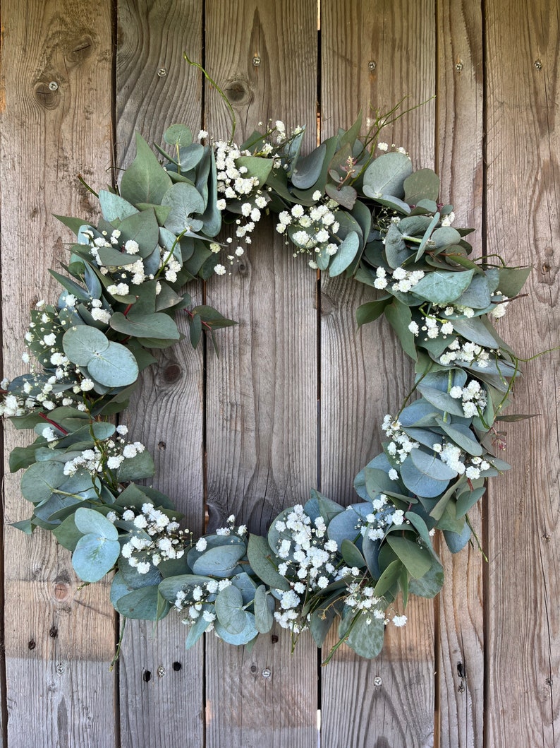 May include: A green eucalyptus and baby's breath wreath hanging on a wooden fence.