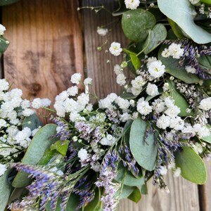 May include: A green eucalyptus wreath with white baby's breath and purple flowers.