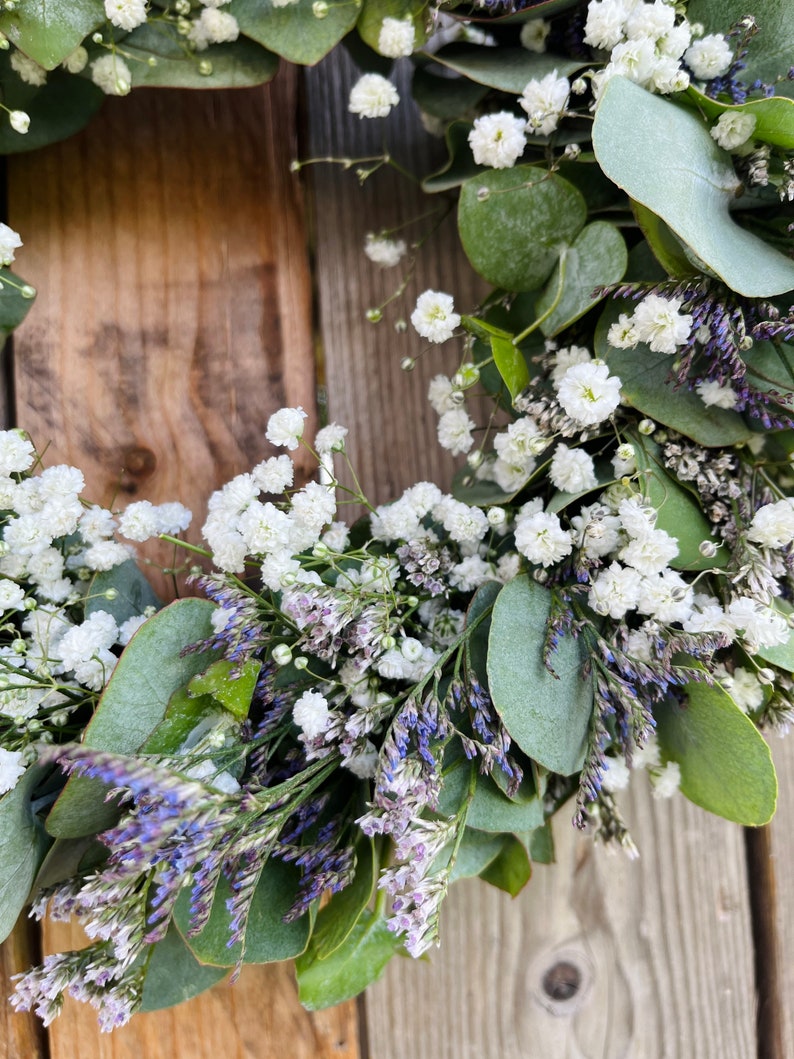 May include: A green eucalyptus wreath with white baby's breath and purple flowers.