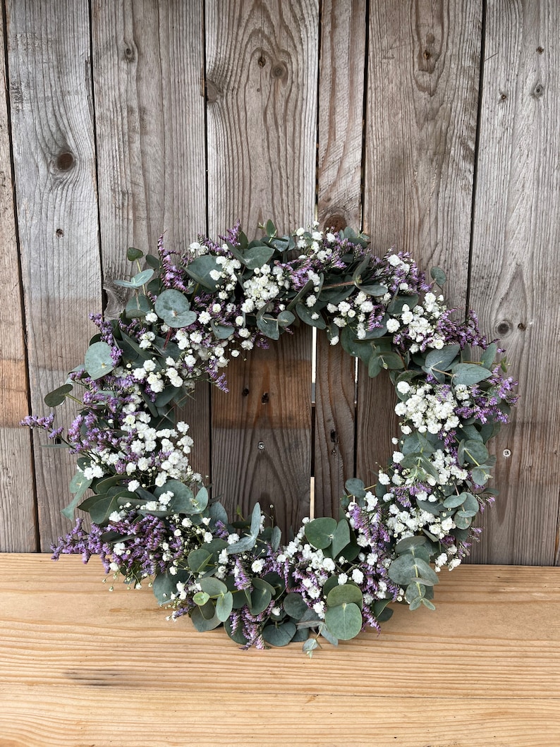 May include: A wreath made of eucalyptus and baby's breath flowers. The wreath is a light green colour with white flowers and purple accents.