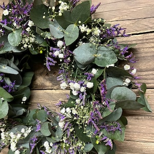 May include: A wreath made of eucalyptus leaves and purple and white flowers. The wreath is hanging on a wooden background.