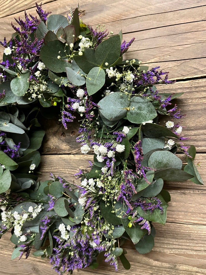 May include: A wreath made of eucalyptus leaves and purple and white flowers. The wreath is hanging on a wooden background.