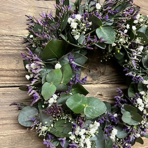 May include: A wreath made of eucalyptus leaves and purple and white flowers. The wreath is hanging on a wooden background.