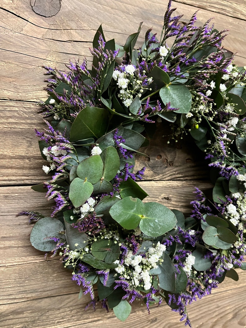 May include: A wreath made of eucalyptus leaves and purple and white flowers. The wreath is hanging on a wooden background.