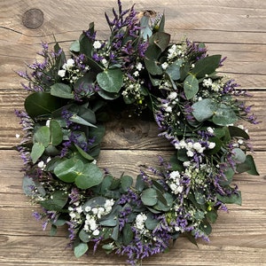 May include: A green eucalyptus and purple flower wreath with white flowers.