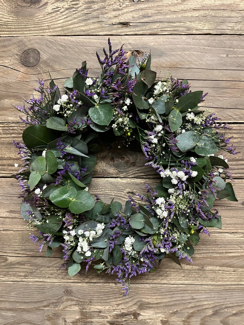 May include: A green eucalyptus and purple flower wreath with white flowers.