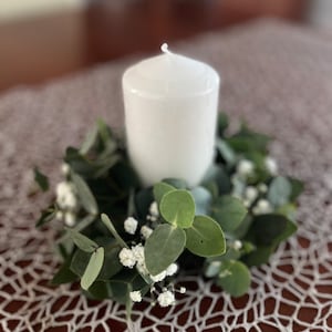 May include: A white pillar candle sits in the centre of a green eucalyptus and white baby's breath wreath.