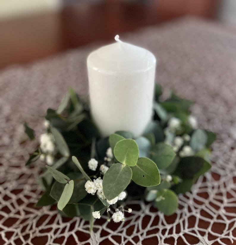 May include: A white pillar candle sits in the centre of a green eucalyptus and white baby's breath wreath.