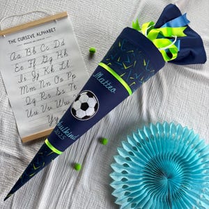 Könnte beinhalten: Ein blauer und grüner Schultüte mit einem Fußball-Design. Die Tüte ist mit einem grünen Band verziert und der Name "Matteo" ist auf der Seite gestickt. Die Tüte trägt auch den Text "Schulkind 2025" auf der Seite.