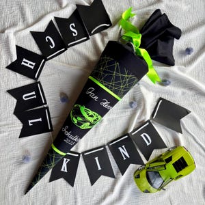 Könnte beinhalten: Ein schwarzer und grüner Schultüte mit einem grünen Band und einem schwarzen und grünen Autodesign. Die Tüte trägt den Text "Jan Hero" und "Schulkind 2023". Es gibt schwarze Flaggen mit weißen Buchstaben, die "Schulkind" um die Tüte herum buchstabieren.
