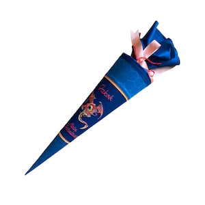 Pode incluir: Um cone escolar azul e dourado com um desenho de drag&atilde;o vermelho. O cone tem o nome "Jakob" e o texto "Mein Schultag" escrito nele. &Eacute; decorado com um la&ccedil;o rosa e vermelho.