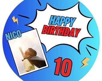May include: A blue and red birthday cake topper with a superhero theme. It features a photo of a child in a cape, a speech bubble with the text "HAPPY BIRTHDAY", and the number "10". The topper also includes a lightning bolt graphic and the name "NICO".