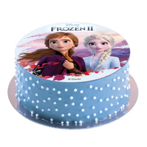 Pode incluir: Um bolo azul e branco com uma imagem comest&iacute;vel redonda de Elsa e Anna de Frozen II da Disney. A imagem tem um fundo de p&ocirc;r do sol rosa e roxo com montanhas ao longe. O texto "Disney FROZEN II" est&aacute; impresso no bolo.